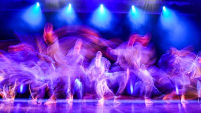 A blurred images of young dancers moving quickly across a large stage with blue lighting from above and purple lighting from the sides