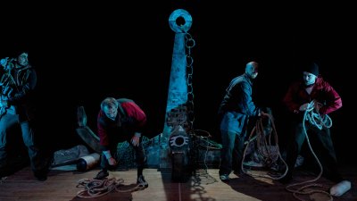 Five performers on stage under a very dim blue light with the large stage set of an anchor on stage in the middle of the group