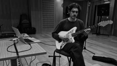 A black and white image of a person sat playing the guitar next to a table with lots of music technical equipment