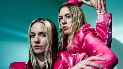 2 female performers wearing electric pink coats dancing against a teal green background