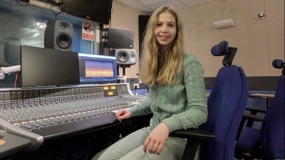 An image of Estella Hurlock sat on a blue chair wearing a green outfit in front of a large audio mixing desk
