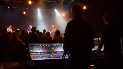 A silhouette image of a sound technician at a mixing desk, a band on stage under colourful theatrical light and an audience in between 