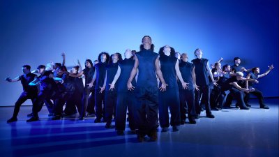 A large ensemble of youth dancers in dark clothing against a striking blue backdrop