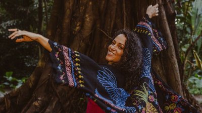 A performer in the woods in front od a tree with the arms in there air looking back over their shoulder towards the camera