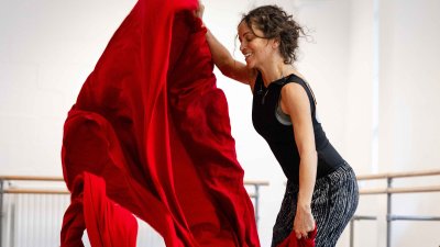 A dancer wearing dark clothing with a large piece of red fabric in a white dance studio