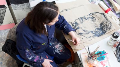 Photo of a young woman sat at a table with a drawing they are working on