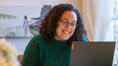 A woman with curly hair and glasses laughing at a laptop