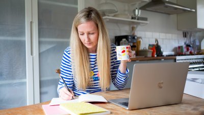 Woman at laptop writing script on paper, holding mug