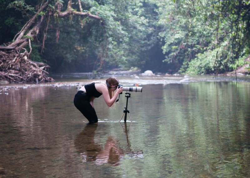 Falmouth University student immersed up to knees in river, bending down to view though camera on tripod.