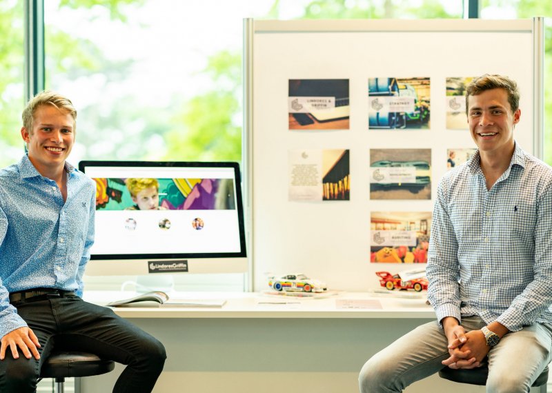 Falmouth University Business students in shirts sat in front of desk with work displayed.