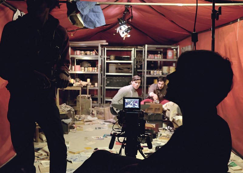Silhouettes of crew on set in red tent, supplies on metal shelves in background, paperwork strewn across the floor.