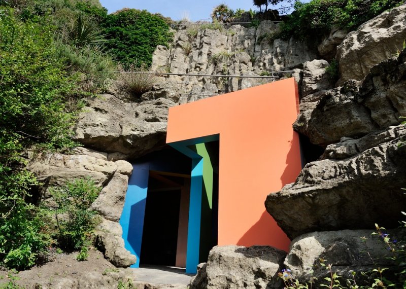 Bright coloured clock building built into the side of a cliff.