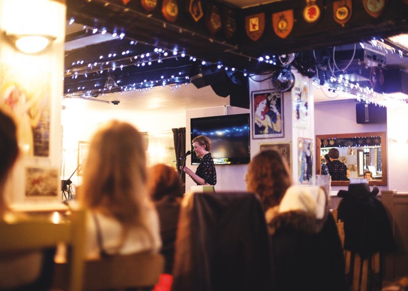 Girl reading from paper in hands in front of audience in pub.