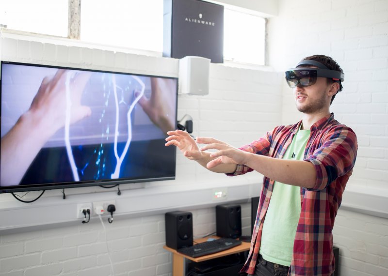 Student wearing virtual reality headset with arms extended in front and images of hands and electricity bolts on screen next to him.