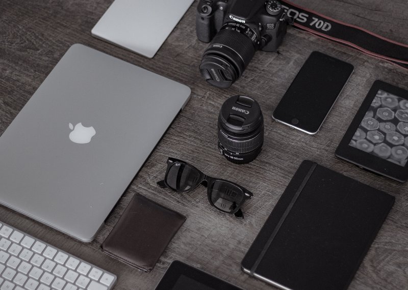 A desk with laptop, keyboard, camera, lens, smartphone.