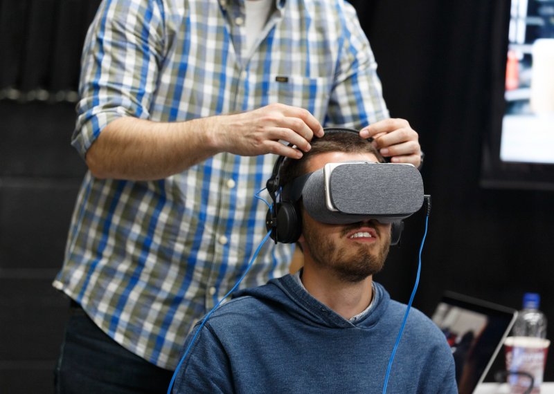 man seated wearing VR headset with another man stood behind him