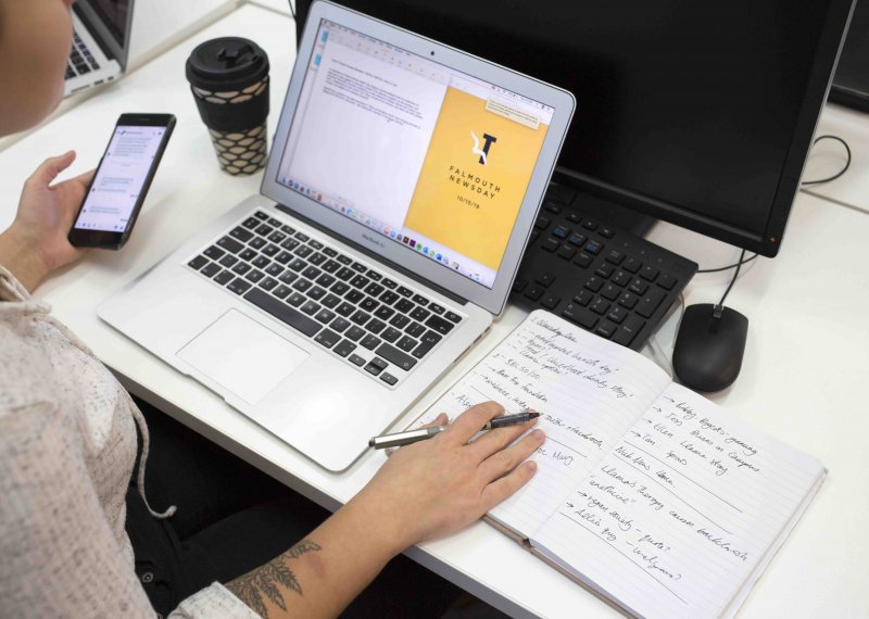 A Journalism student working at a laptop and holding a phone