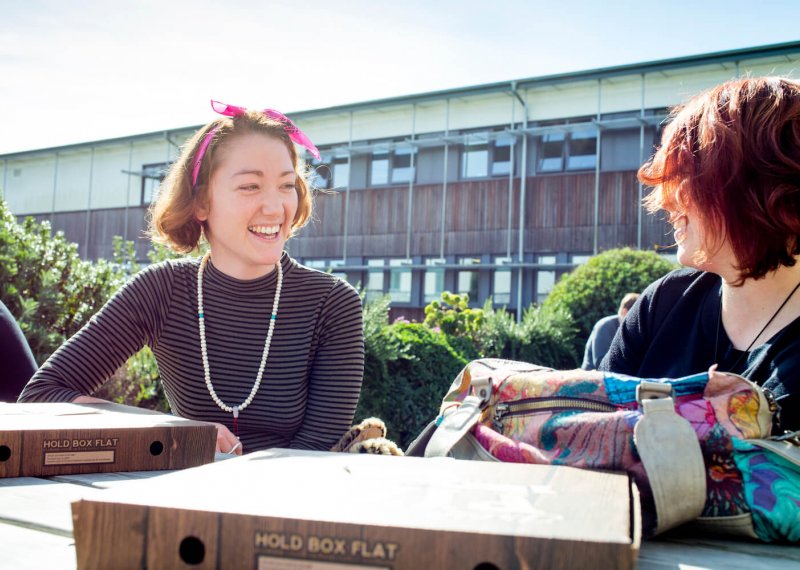 Two female students smiling sat at a picnic bench on Penryn Campus.