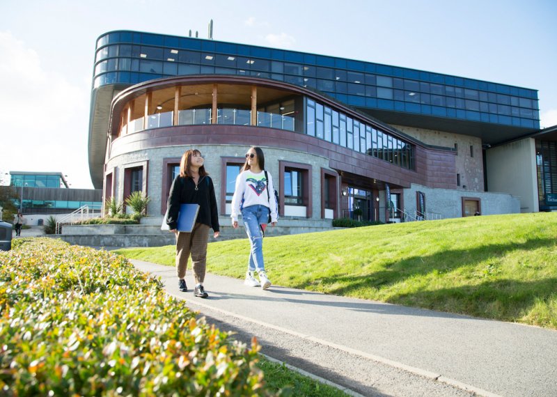 Two students walking past a large building on Penryn Campus