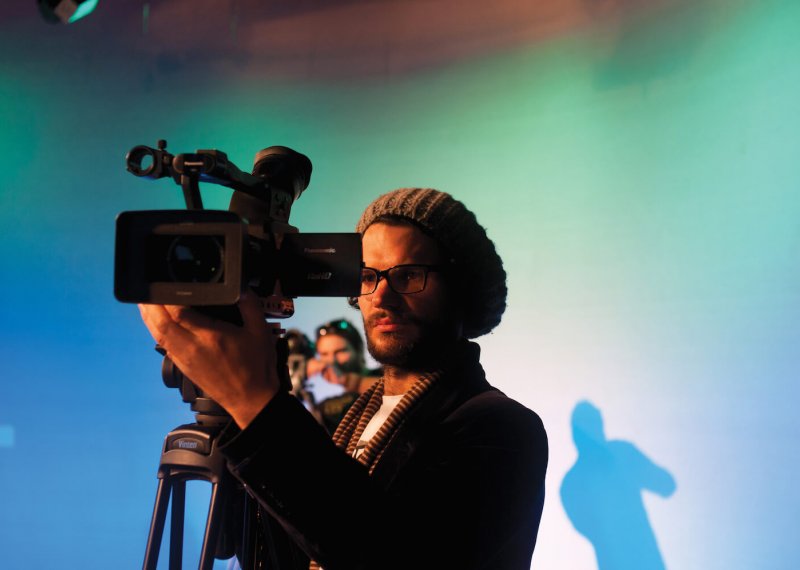 A male student wearing a hat and operating a film camera in a blue studio