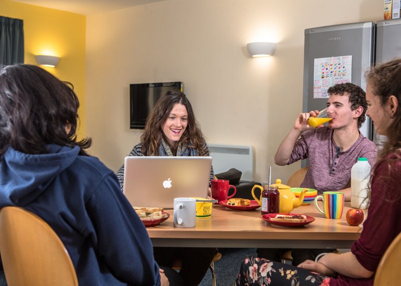 Students around kitchen table drinking orange juice and looking at laptop.