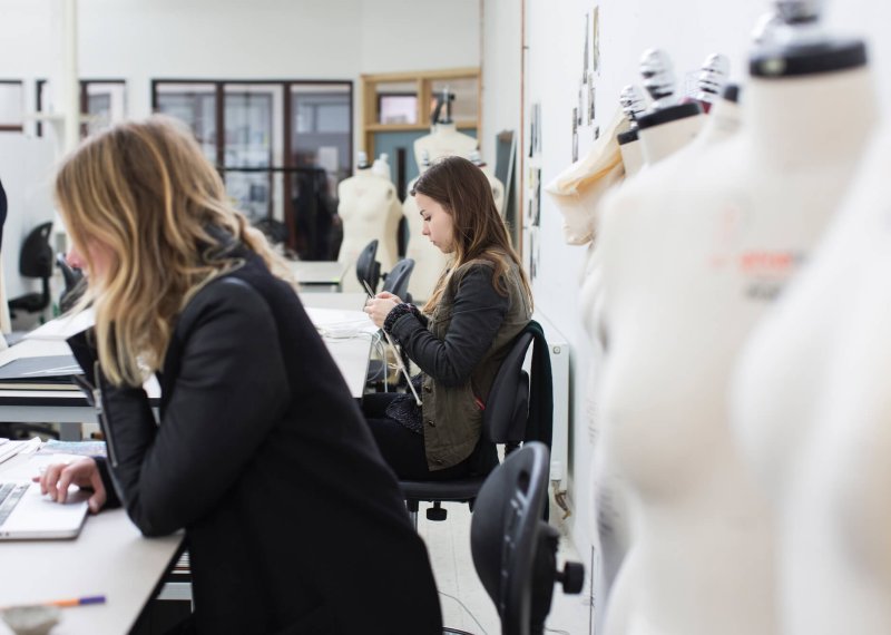 Two students in fashion studio, one on laptop and one knitting.