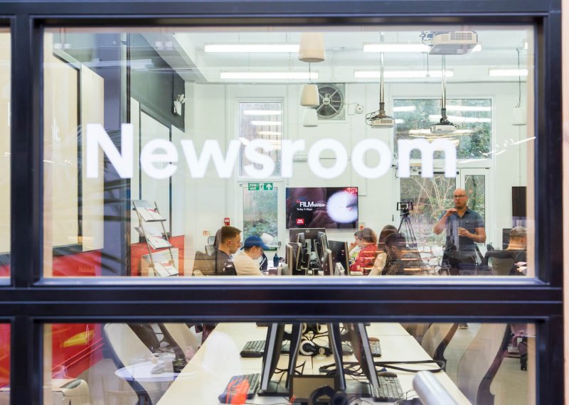 Through the window of the newsroom with lecturer talking to journalism students.