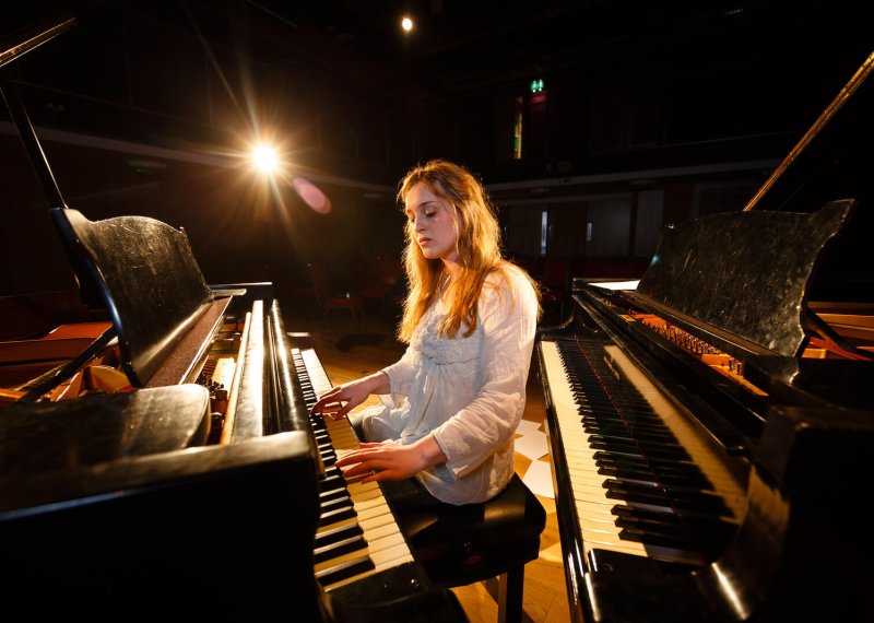 Female student sat between two pianos and playing one of them, sheet music on floor.