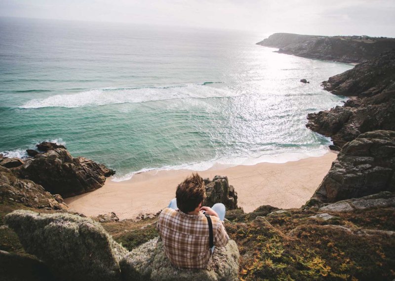 Man sat on the edge of cliff looking out to sun reflections on the water and coastline.