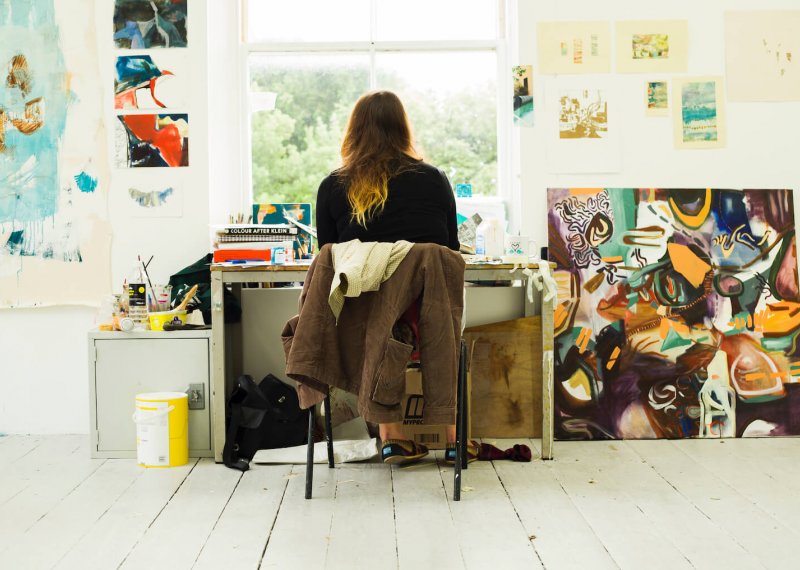 Back of student sitting at desk in front of window surrounded by artwork and paintings.
