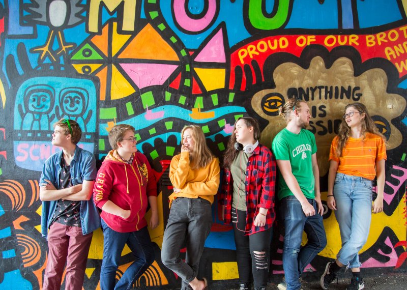 Students hanging out in a line in front of brightly graffitied wall in Falmouth.