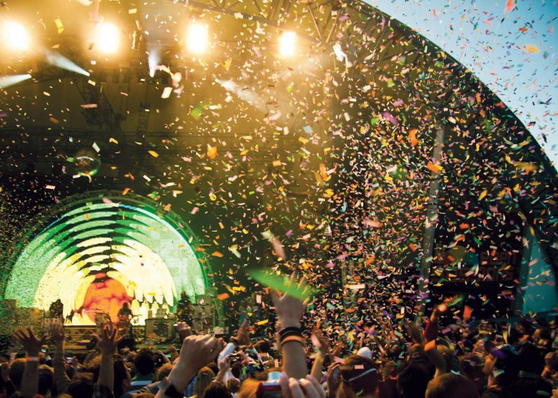 A large crowd with their hands in the air watching a music performance on a stage outside