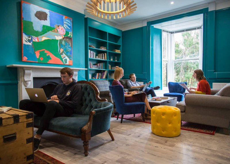 Falmouth University students sitting on armchairs and sofas in a teal room with wooden floor