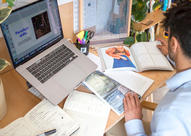 A man sat at a computer with fashion-related books and paper