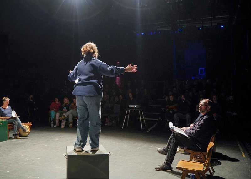 Performer with their back to the camera on stage under a spotlight performing to an audience