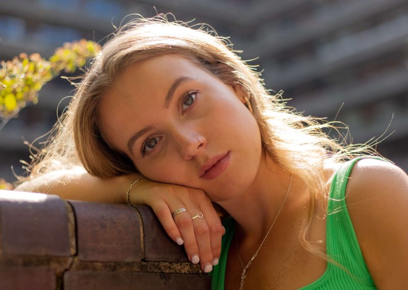 Graduate Eva Gadd wearing a bright green tank top and leaning against a red brick wall, with large block of flats in the background.