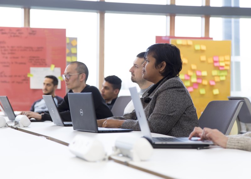 A group of people sat with laptops on a table, board of post-it notes behind them