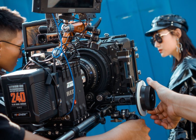 A man controlling a RED camera filming a women wearing sunglasses, a leather hat and jacket