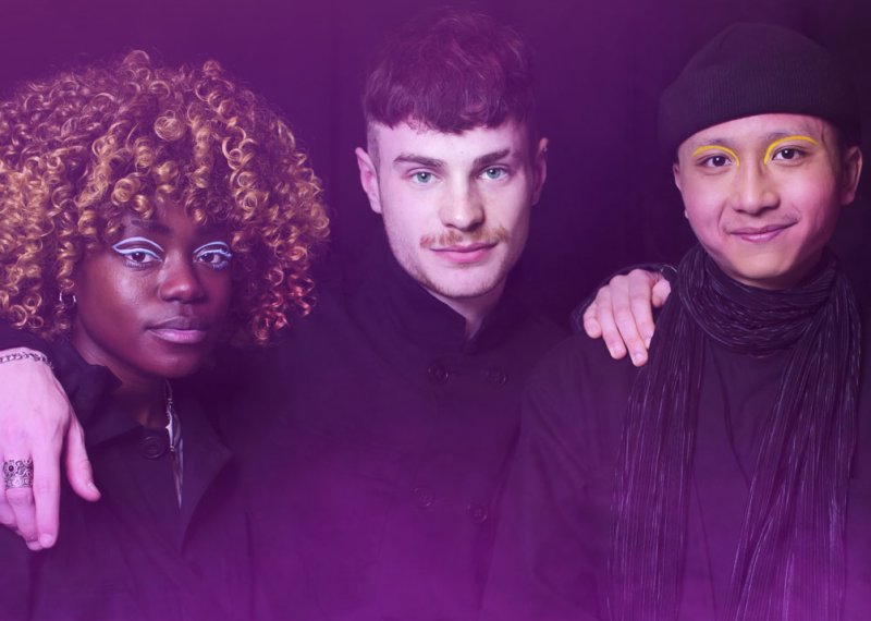 Photo of four student with their arms around each other's shoulders. Background is black with a pink haze.
