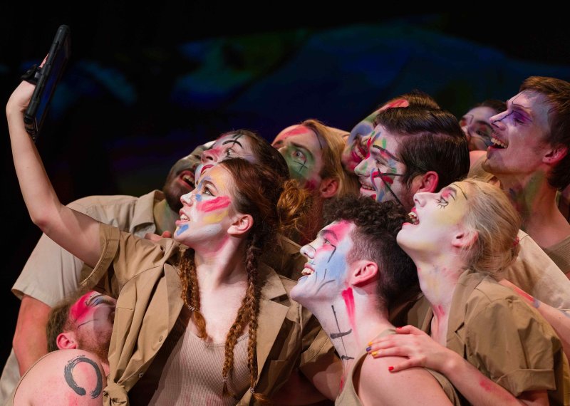 A group of actors with painted faces look towards a hand-held tablet 