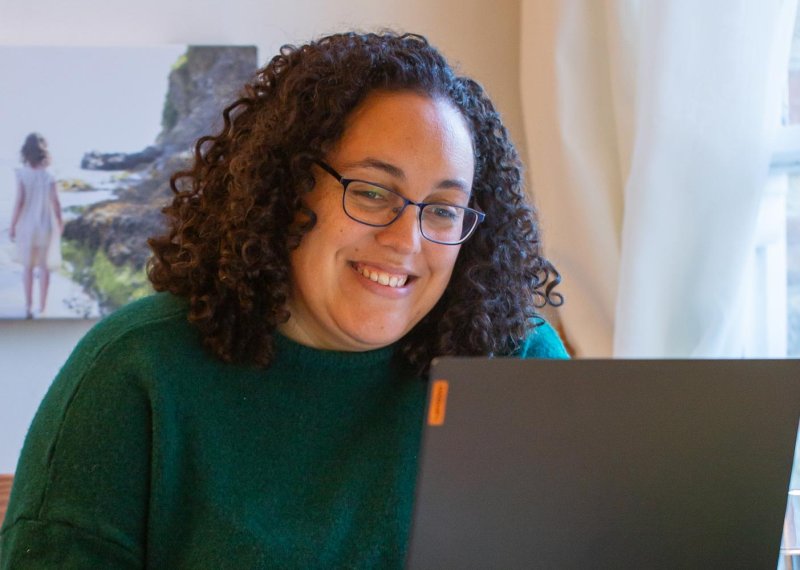 A woman with curly hair and glasses laughing at a laptop