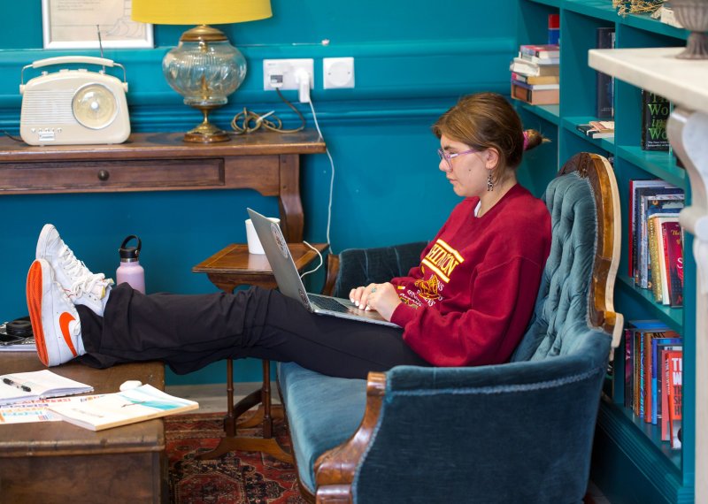 Girl wearing red jumper and pink glasses sits on a blue sofa with her feet up on a coffee table while working on a laptop
