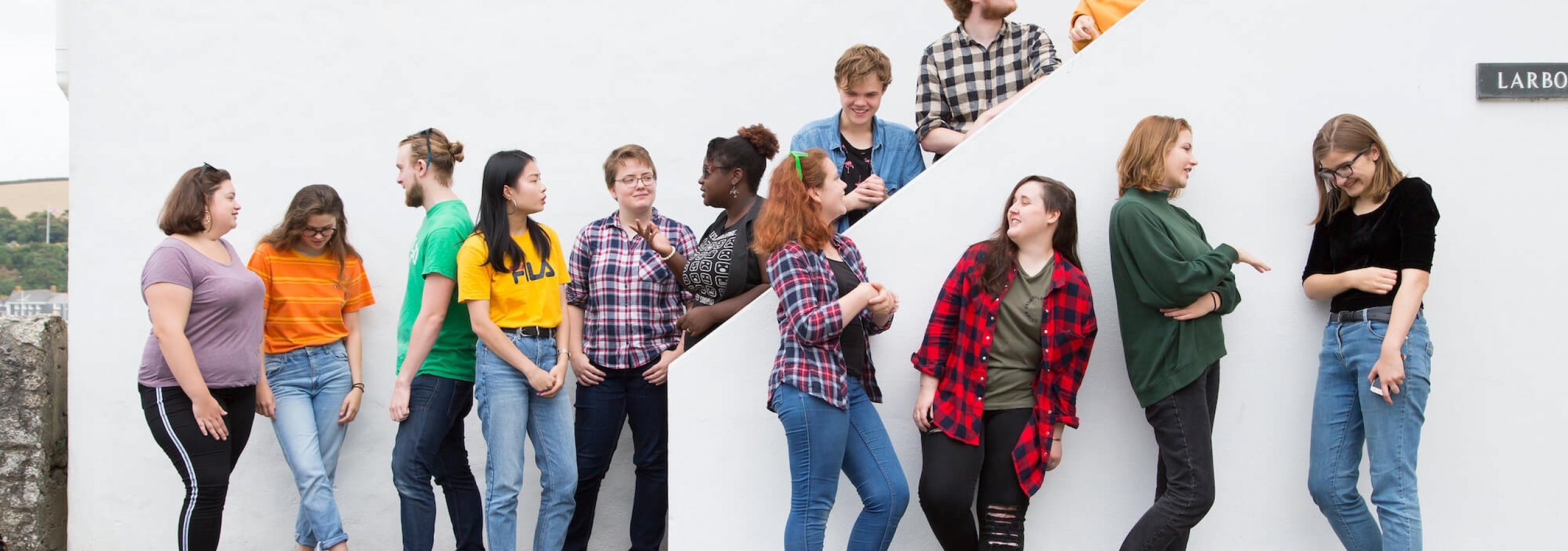 13 Falmouth University students in different coloured tops stood against a white wall and on a white staircase.
