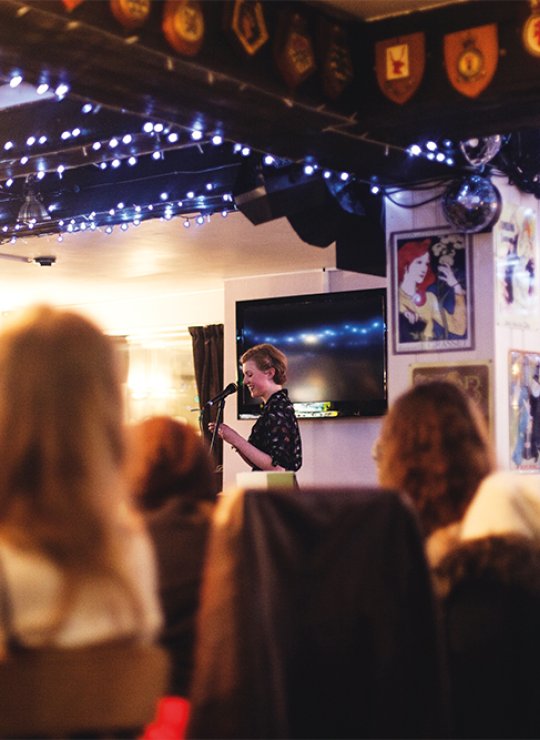 Student speaking into microphone at event in pub surrounded by blue fairy lights.