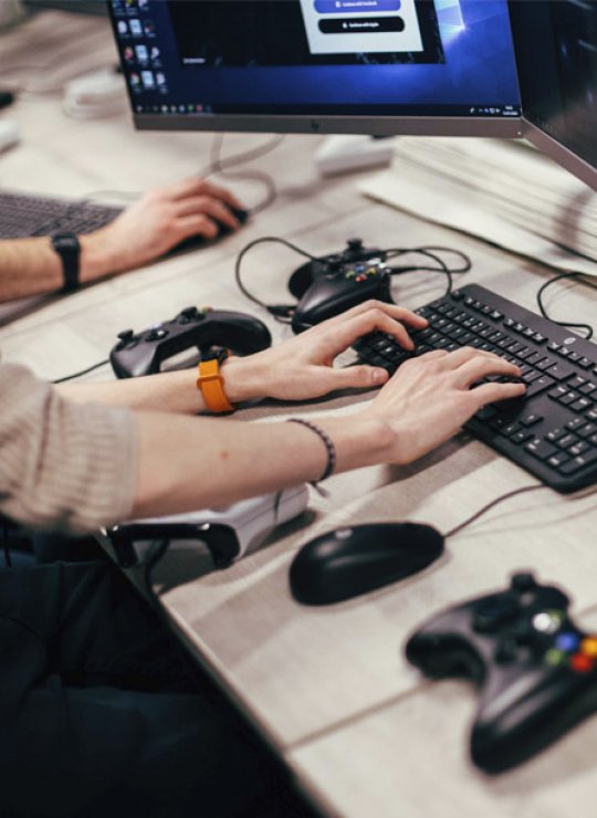 Hands typing on a keyboard with playstation controllers on the table