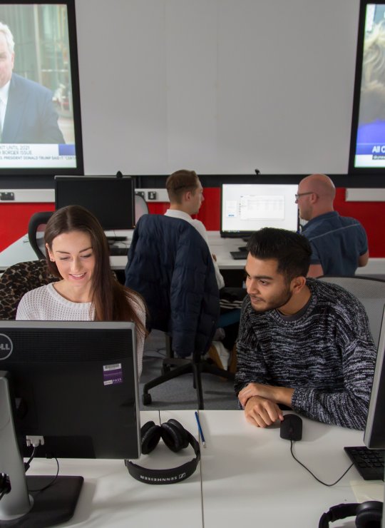 Students working together in newsroom, news playing on screens behind them.