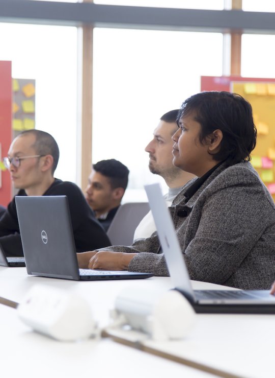 A group of people sat with laptops on a table, board of post-it notes behind them