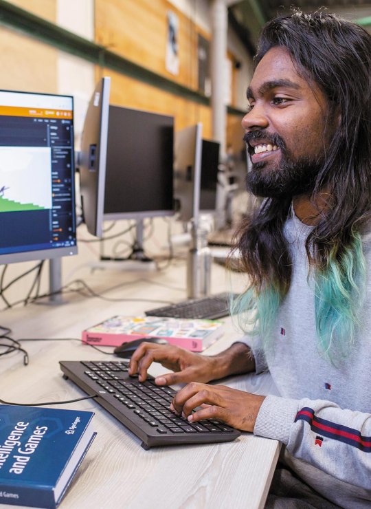 A man typing at a computer with an artificial intelligence for games book on the table