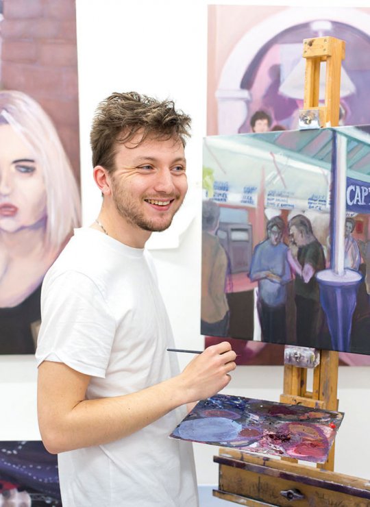 Student stood in front of paintings, smiling and holding paintbrush
