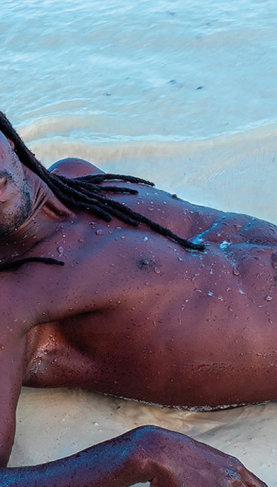 A man with dreadlocks and wearing black shorts, lying on the beach shore line